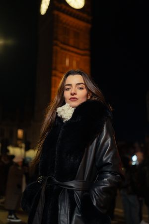 A person in a black leather coat with fur collar poses on a city street at night under illuminated architecture.