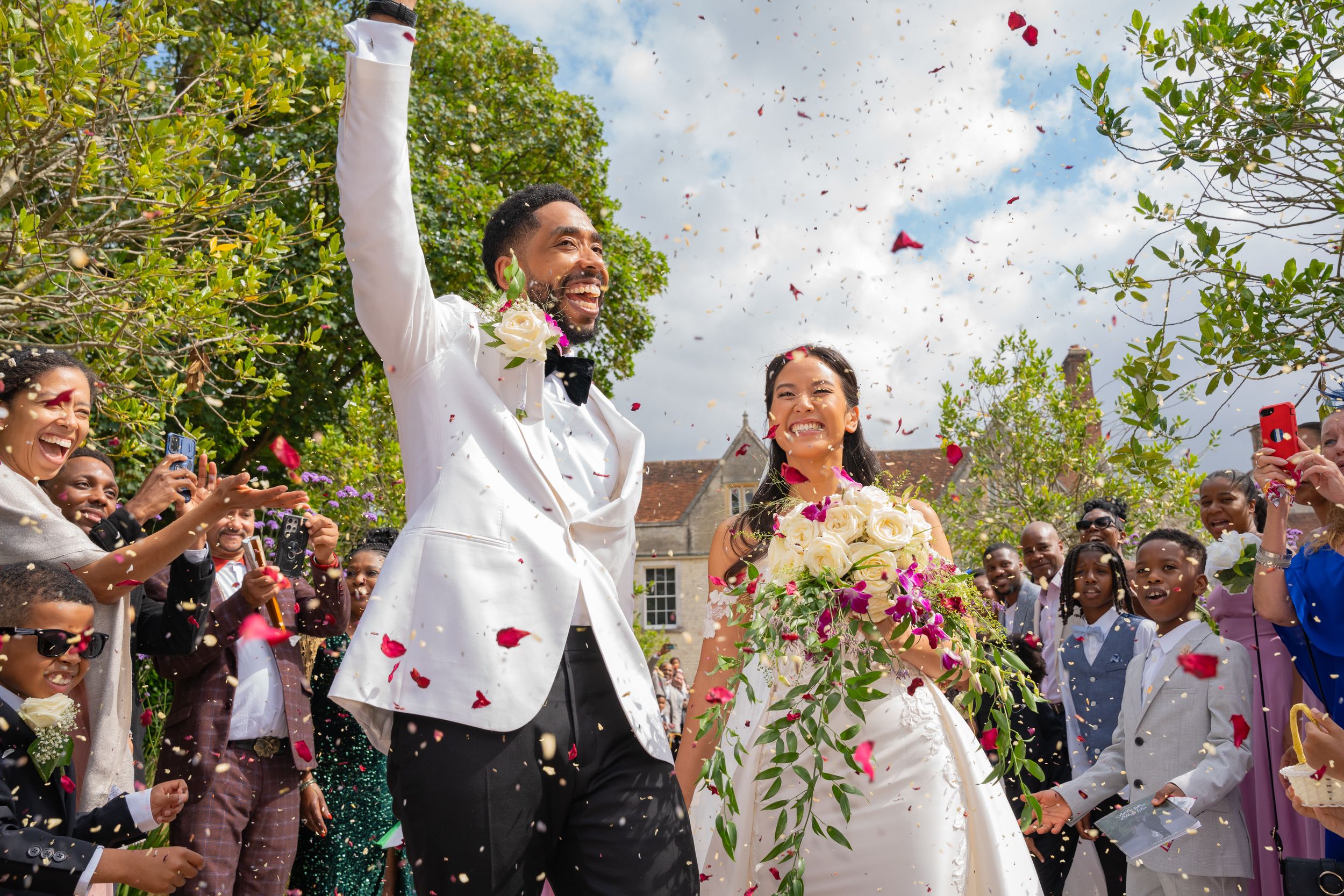Wedding celebration moment with confetti being thrown as newlyweds celebrate outdoors under trees on sunny day.
