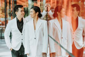 Newlywed couple in matching white outfits pose against mirrored surface in urban setting. Wedding portraits session.