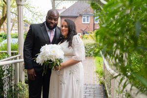 A newlywed couple stands together on a garden path holding a white floral bouquet near a brick house. Registry civil ceremony.