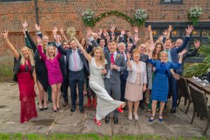 A cheerful wedding party group poses together outside a brick building with raised arms and celebratory gestures.