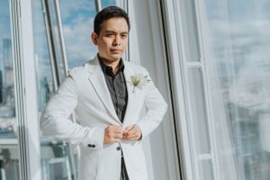Groom in white suit jacket with black shirt and boutonniere poses near window in the Shard, Shangri-La Hotel.