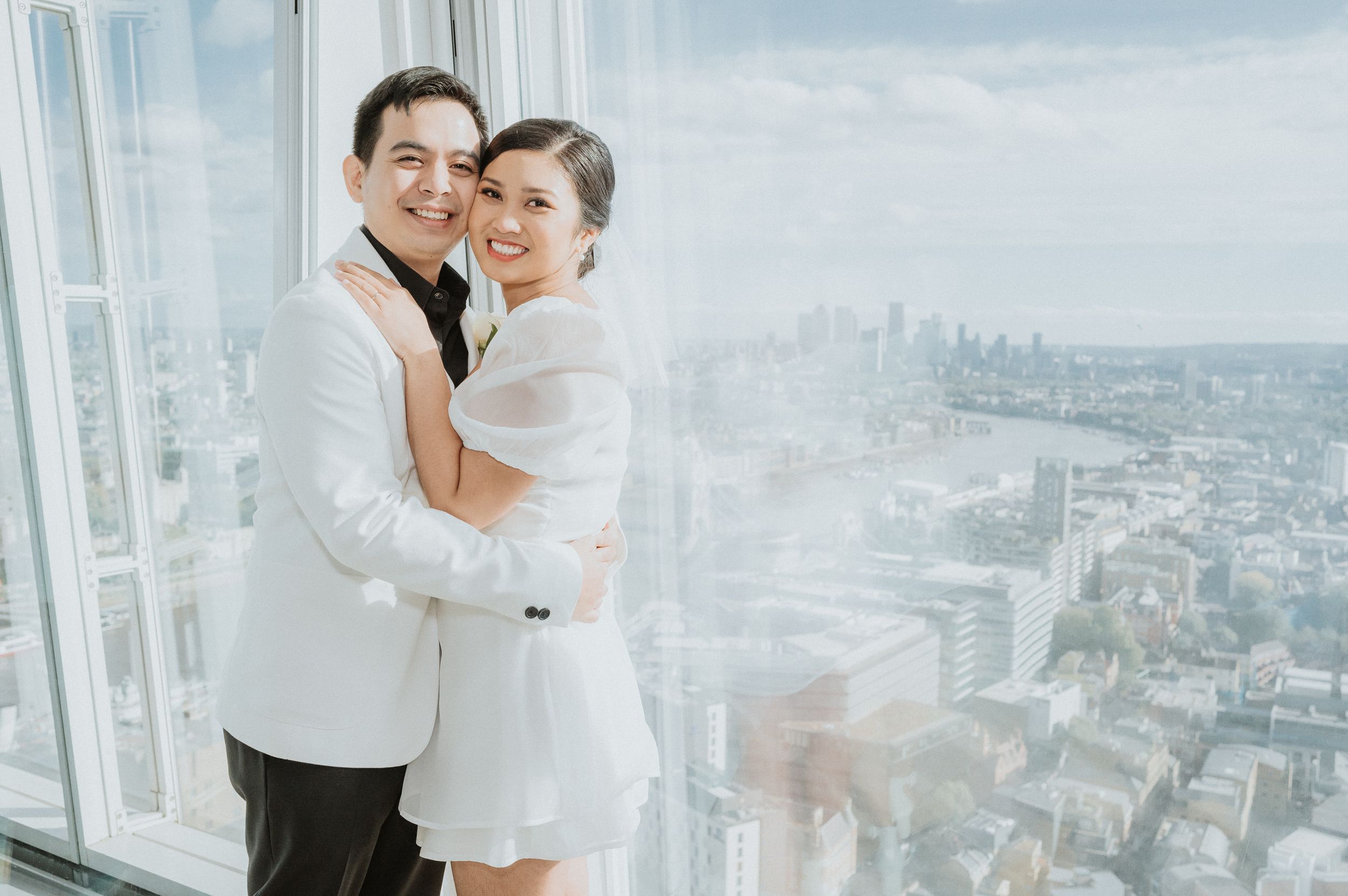 Happy newlywed couple embracing in formal attire against cityscape backdrop.