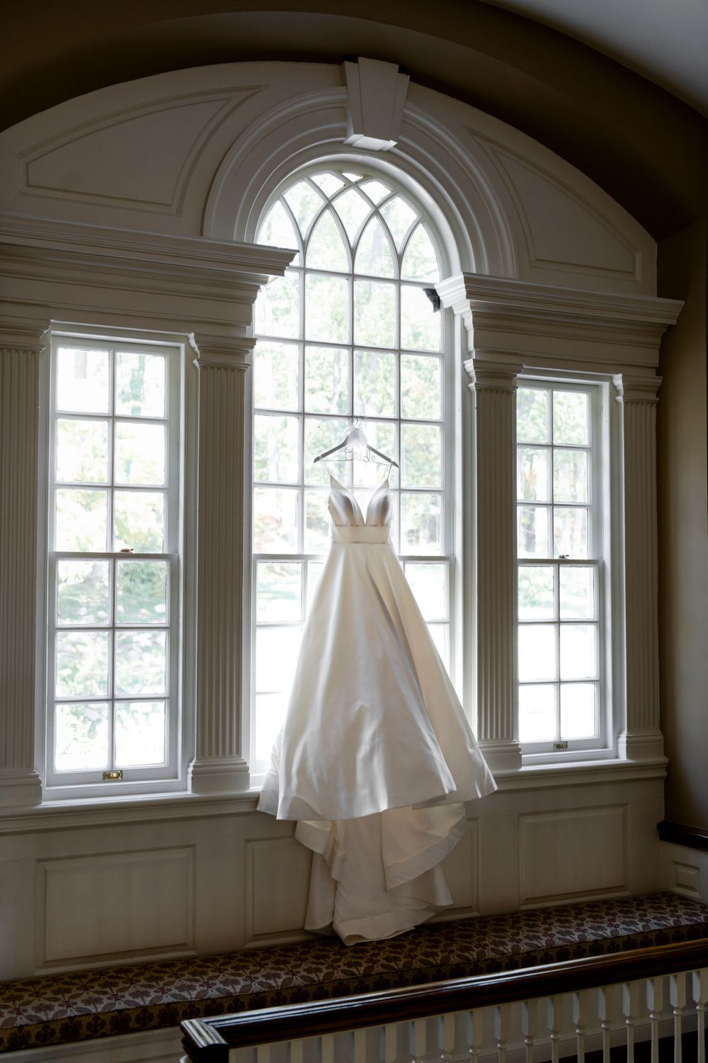 White wedding dress hanging in front of an elegant arched window with natural light streaming through architectural details.