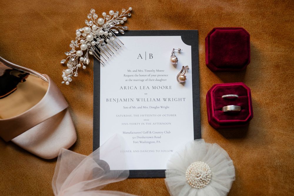 Wedding invitation flatlay with velvet ring box, crystal hair pin, and tulle detail on brown leather background.