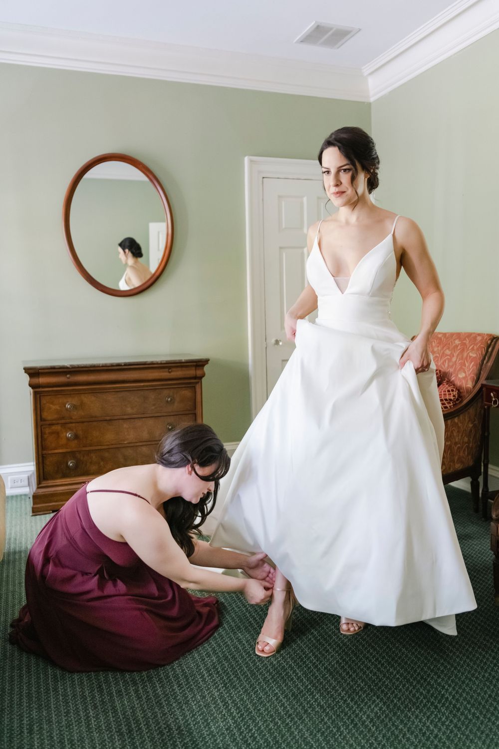 Wedding preparation scene showing a bridesmaid in burgundy helping adjust the bride's shoes in a room with green carpet.