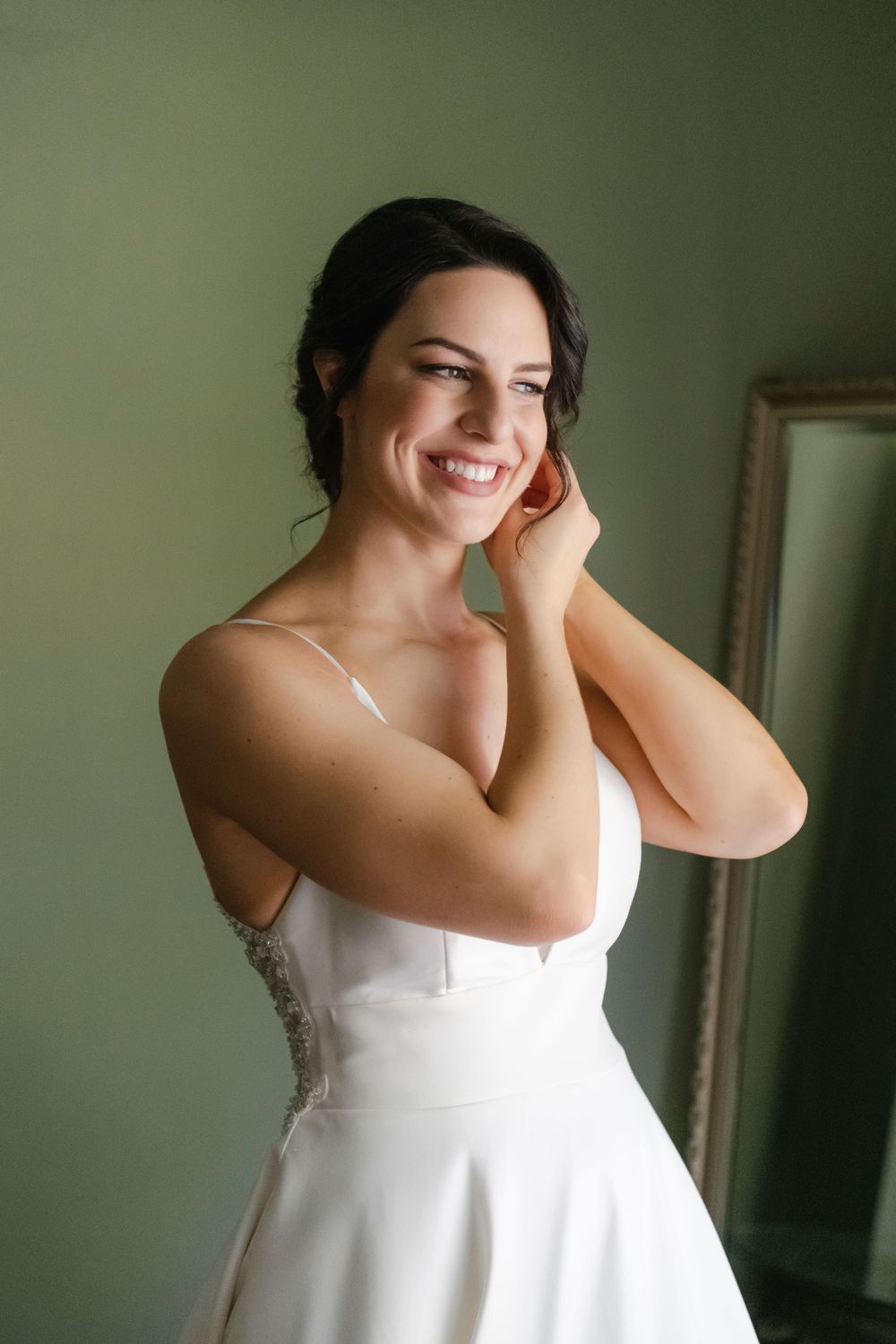 A joyful candid portrait showing the bride putting in her earrings while naturally smiling in soft window light.