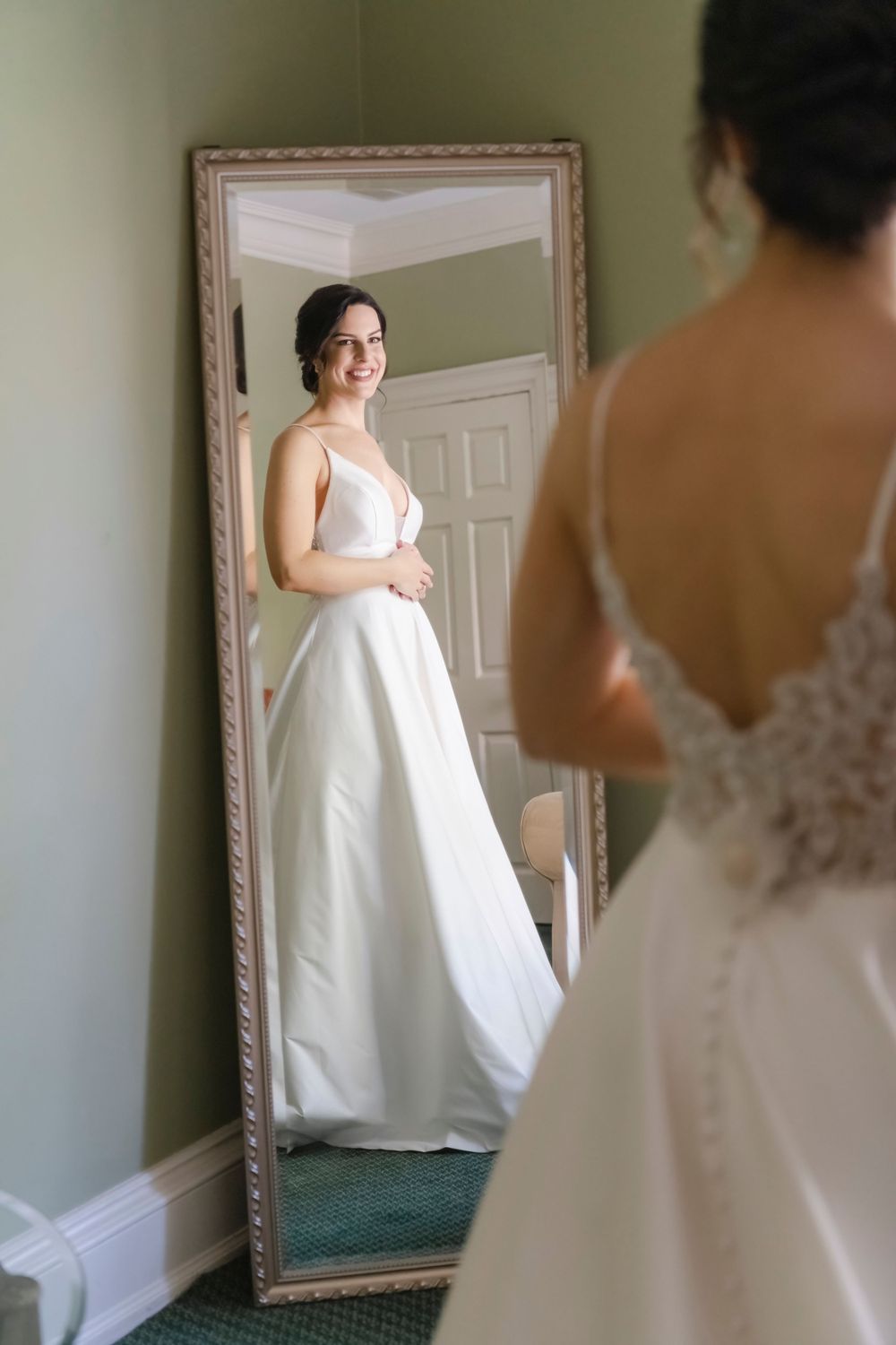 A bride in an elegant white wedding dress looks at her reflection in a full-length ornate framed mirror.