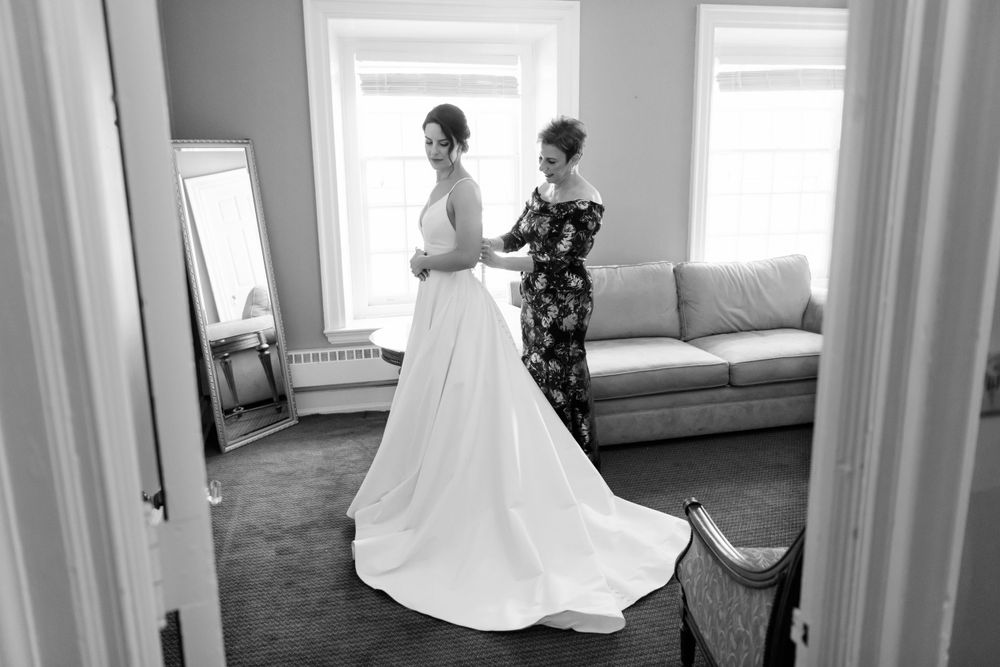 Black and white image shows a classic bridal scene with dress fitting and preparation in an elegant room with natural light.