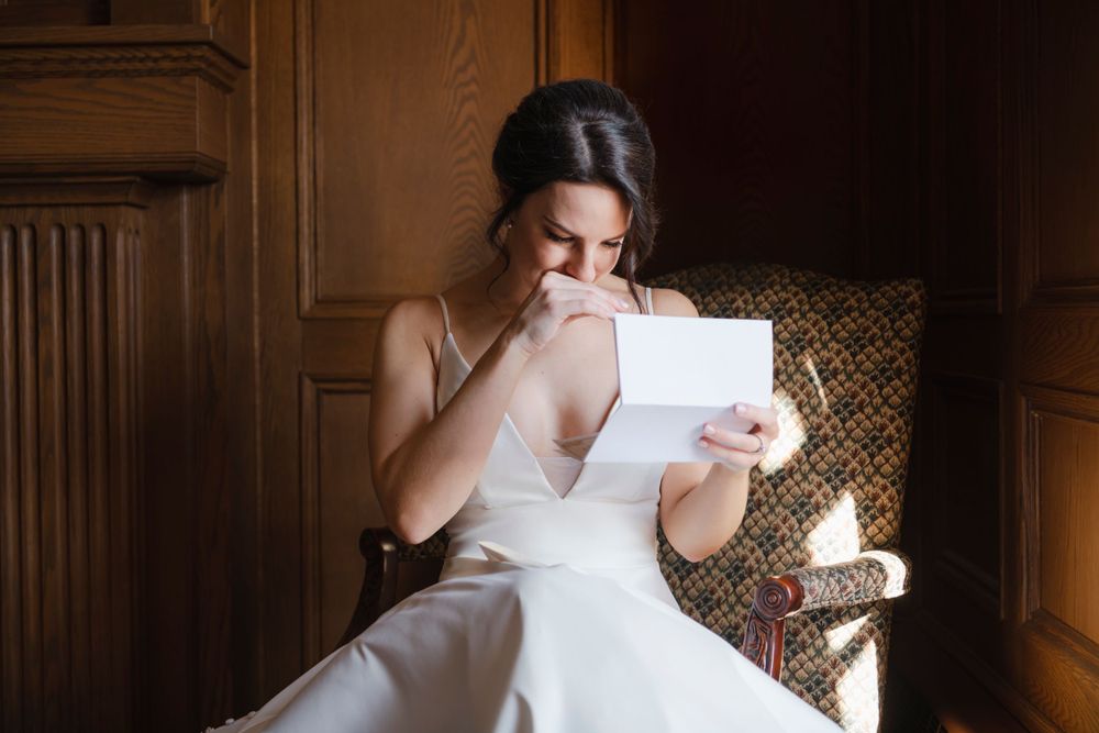 A bride in a white wedding dress reads a letter while sitting in an elegant wooden-paneled room.