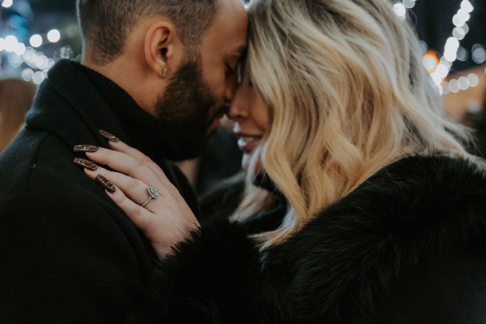 A close-up of a couple touching foreheads in a loving moment, the woman's ring and dark manicure visible in soft light.