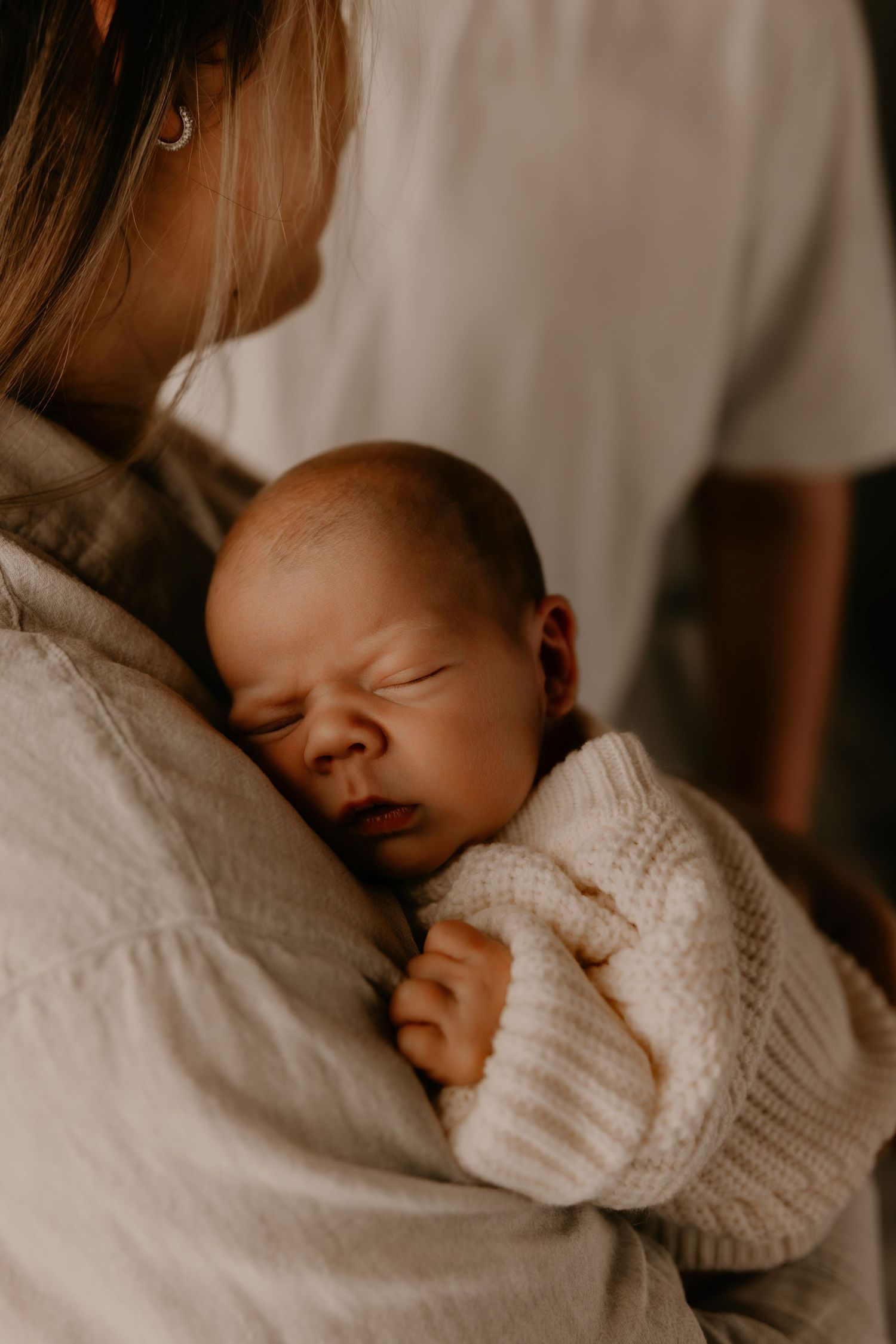 A newborn baby sleeping peacefully in a cream knit sweater, cradled in a mother's arms.