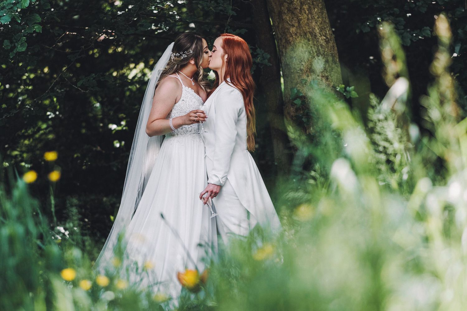 Two brides in white wedding dresses embrace in a wildflower garden.