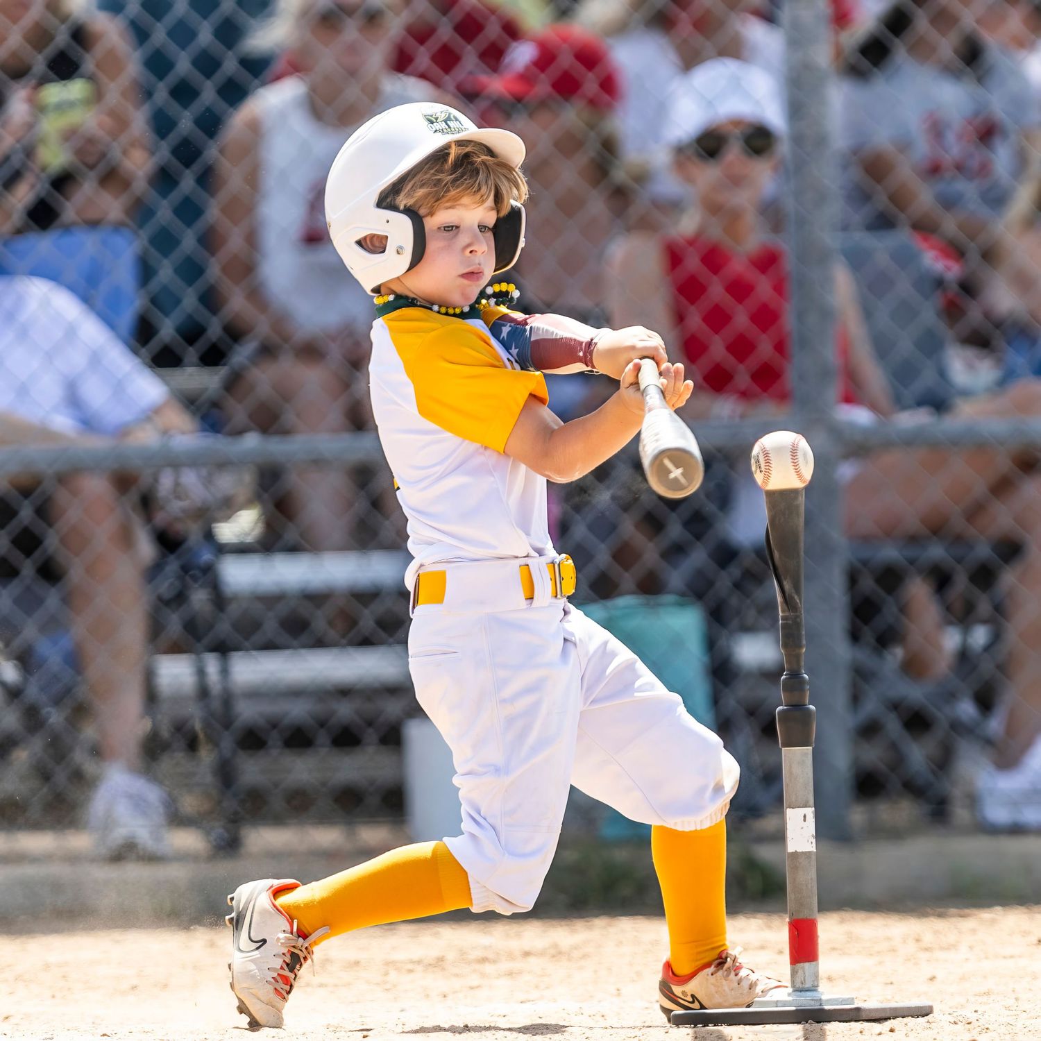 Young boy in white and yellow baseball uniform swinging bat at tee ball during a little league game.