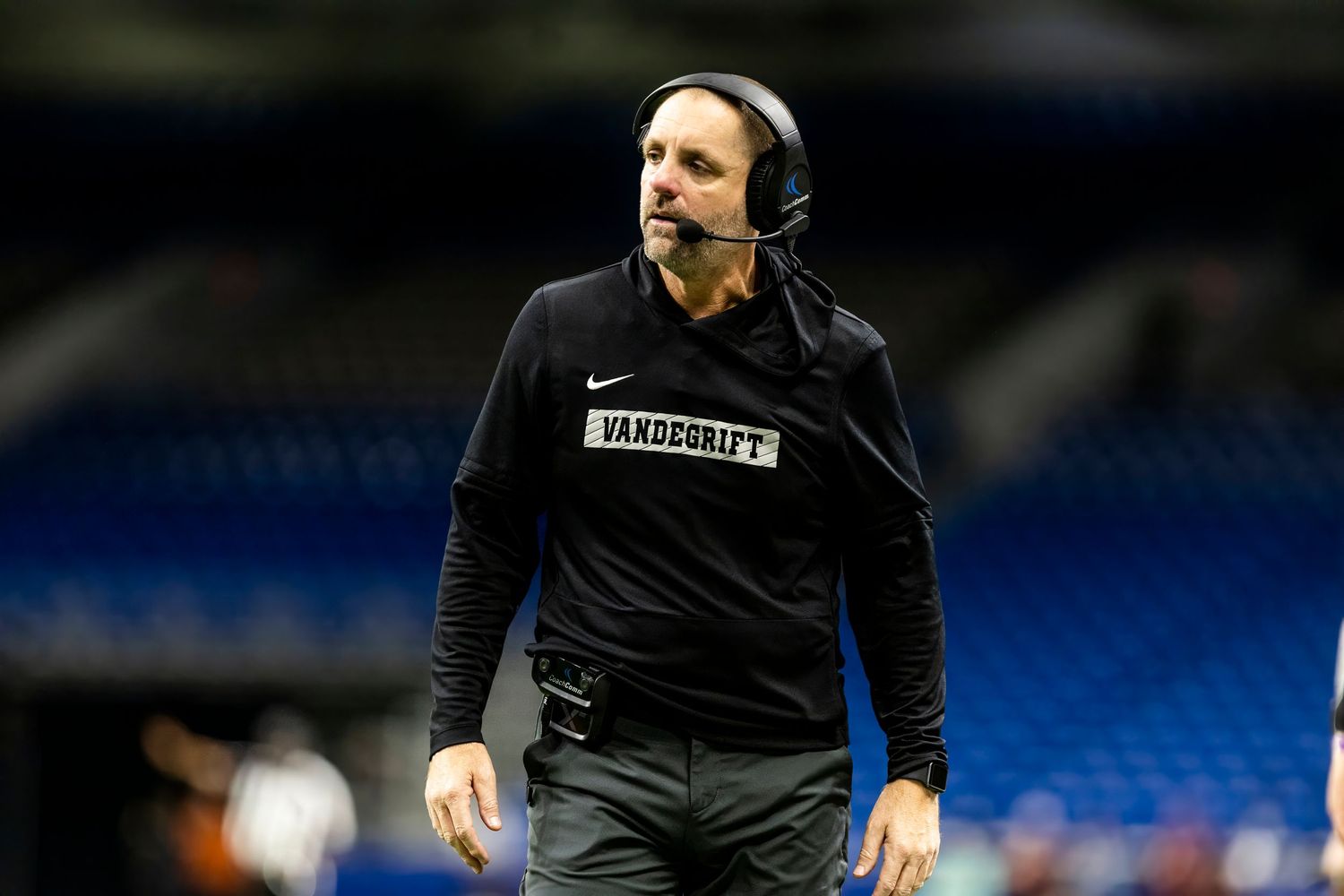 Vanderbilt football coach wearing headset and black Nike gear on the sideline during an indoor stadium game.