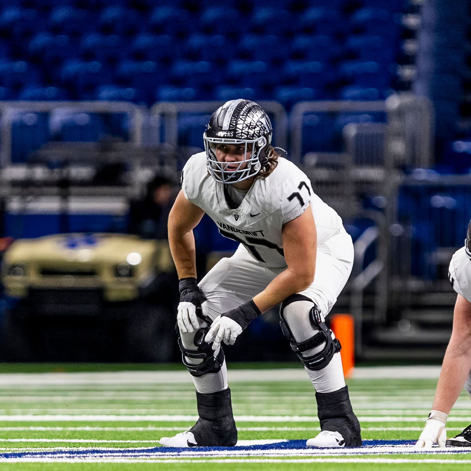 Football player in gray uniform and helmet crouches in offensive line stance on indoor stadium field.