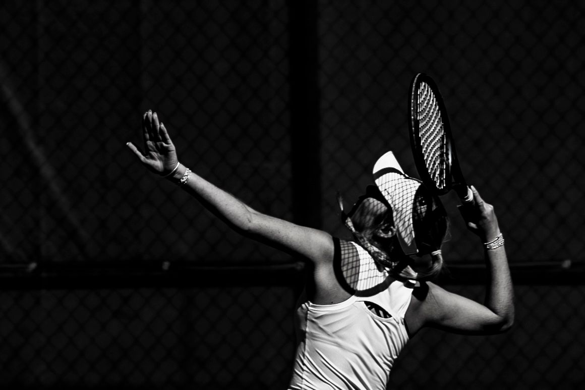 Black and white photo of a female tennis player serving, racket raised overhead on a dark court.