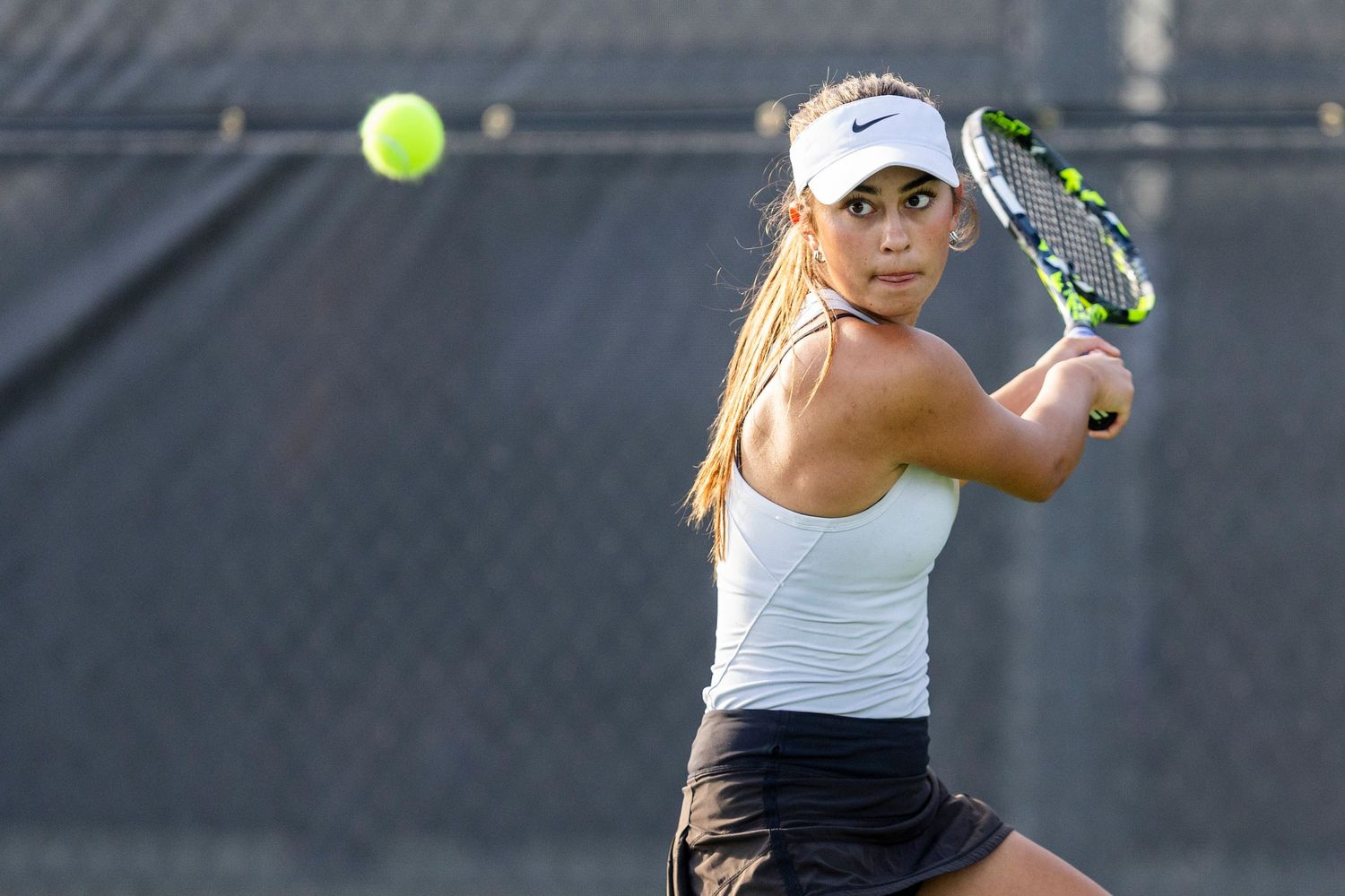 Female tennis player in white top and visor executing a backhand shot on an outdoor court.