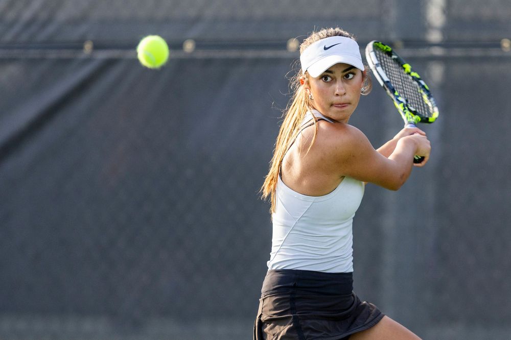 Female tennis player in white top and visor executing a backhand shot on an outdoor court.