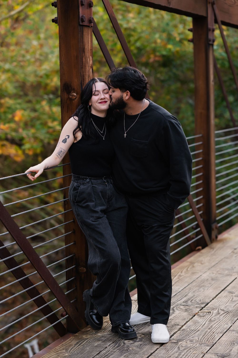 Couple dressed in black on truss bridge during couples session. Girl leaning on bracing with a smile while guy leaning in to kiss her cheek