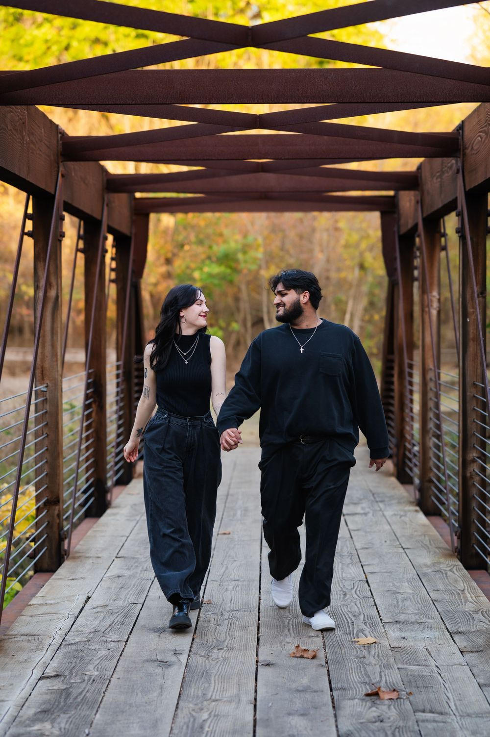 Fashionable couple in black holding hands, looking at each other walking through truss bridge; bright, warm nature scenery in the background