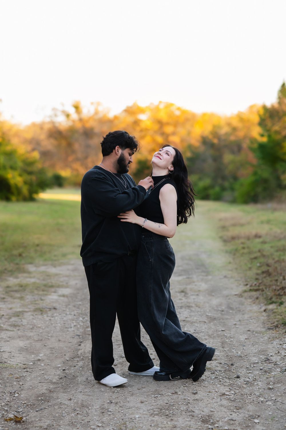 Couple dressed in black embracing lovingly with girls head thrown back and smiling while he adjusts her necklace during outdoor session