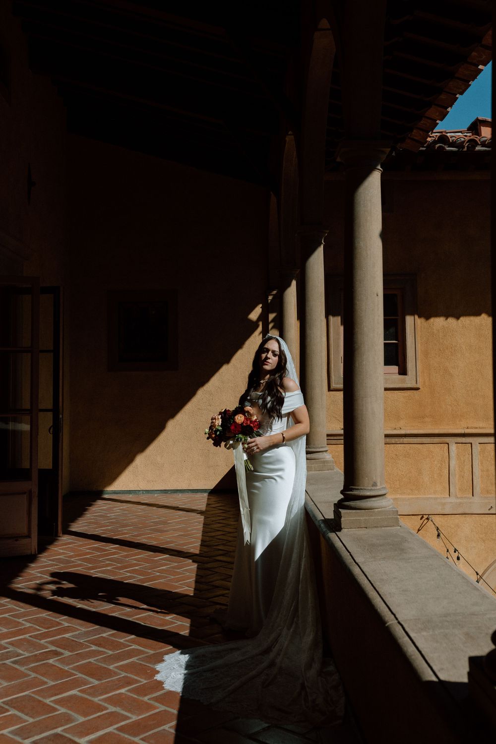 A bride in a white gown stands in dramatic shadows between elegant stone columns on a brick patio walkway.