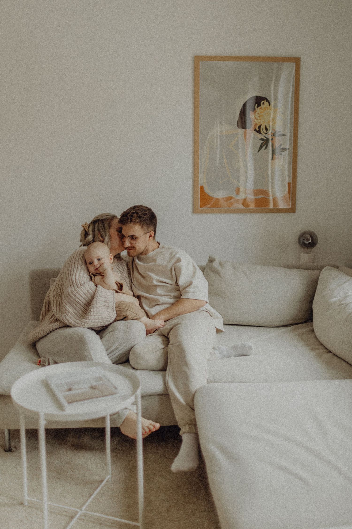 A cozy couple in cream sweaters share an intimate moment on a white couch with minimalist decor in the background.