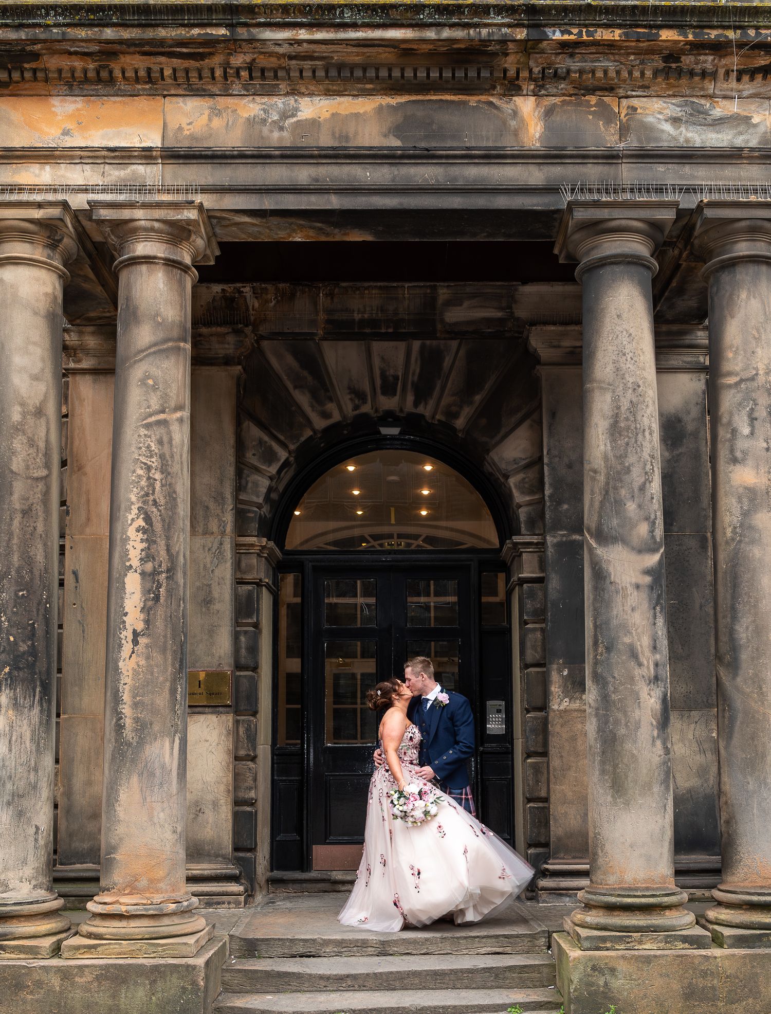 Wedding couple poses under classical stone columns at historic city building entrance.