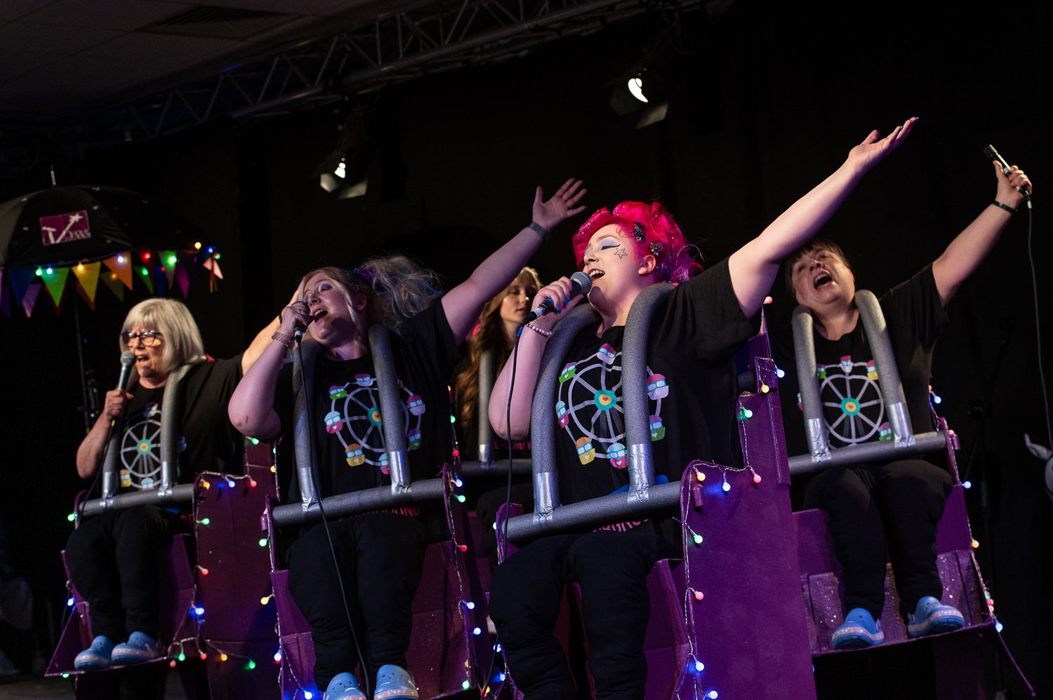 Concert performers energetically dance and move across a dimly lit stage with purple lighting and colorful effects in background.