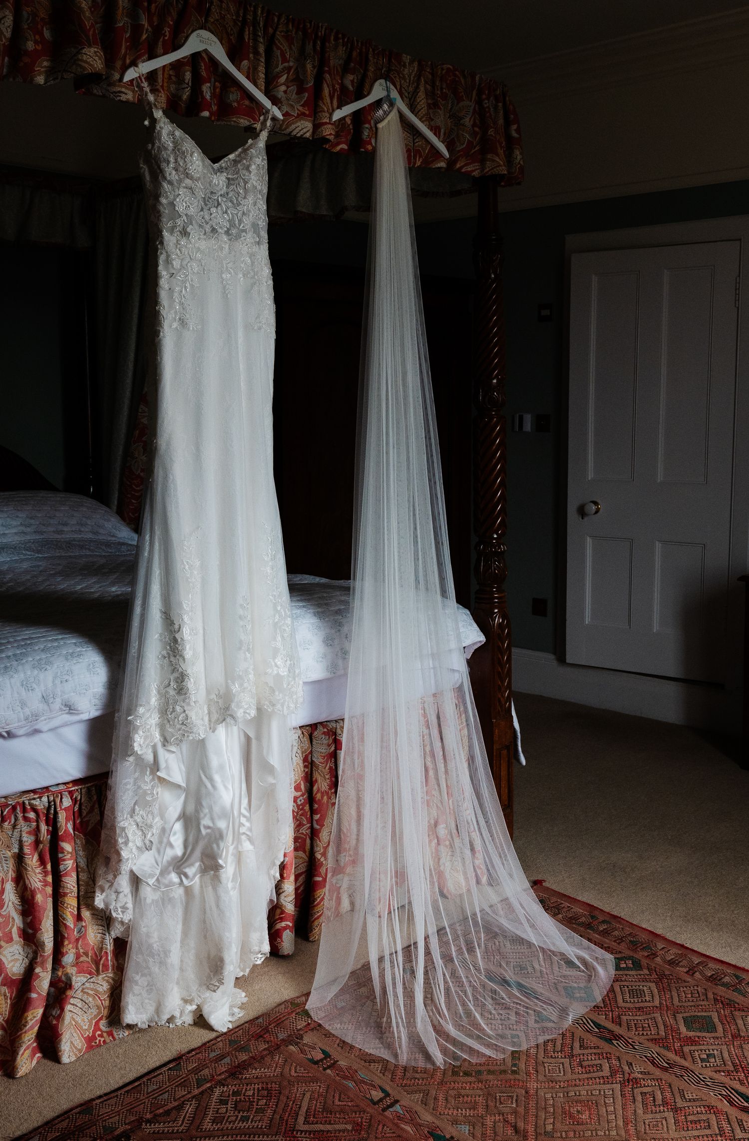 A white lace wedding dress and long sheer veil hanging on decorative hangers beside a canopy bed.