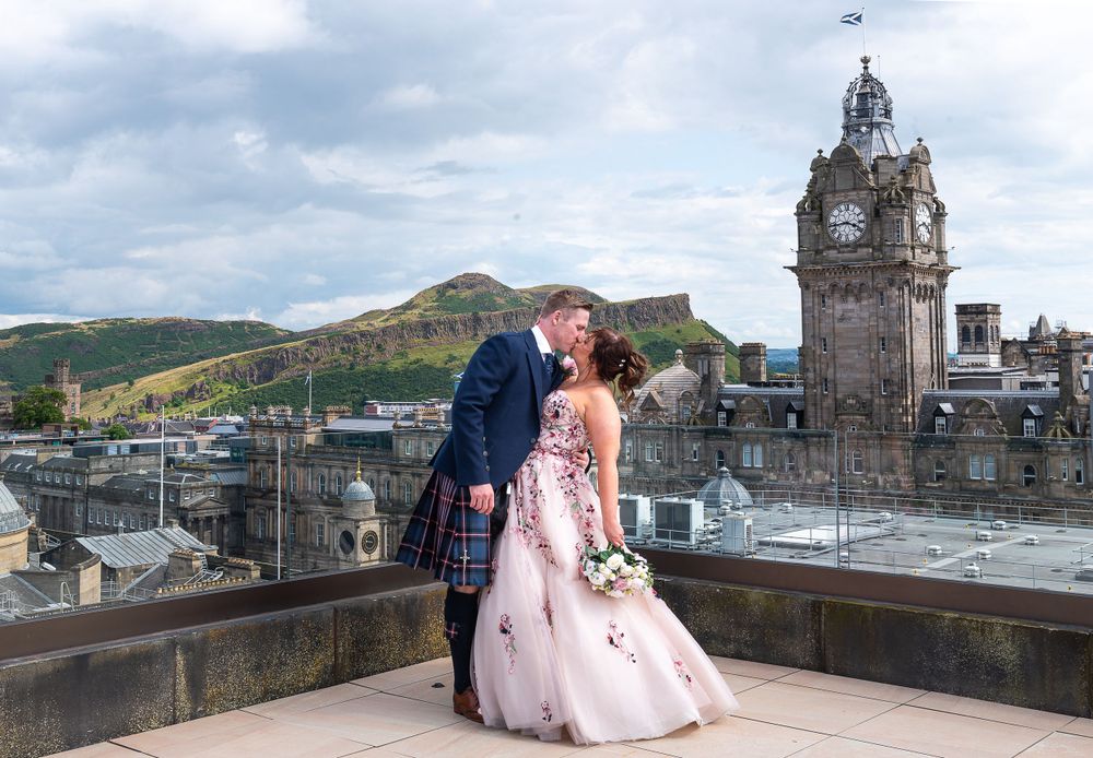 Couple pose on rooftop with historic clock tower and Edinburgh skyline in background.