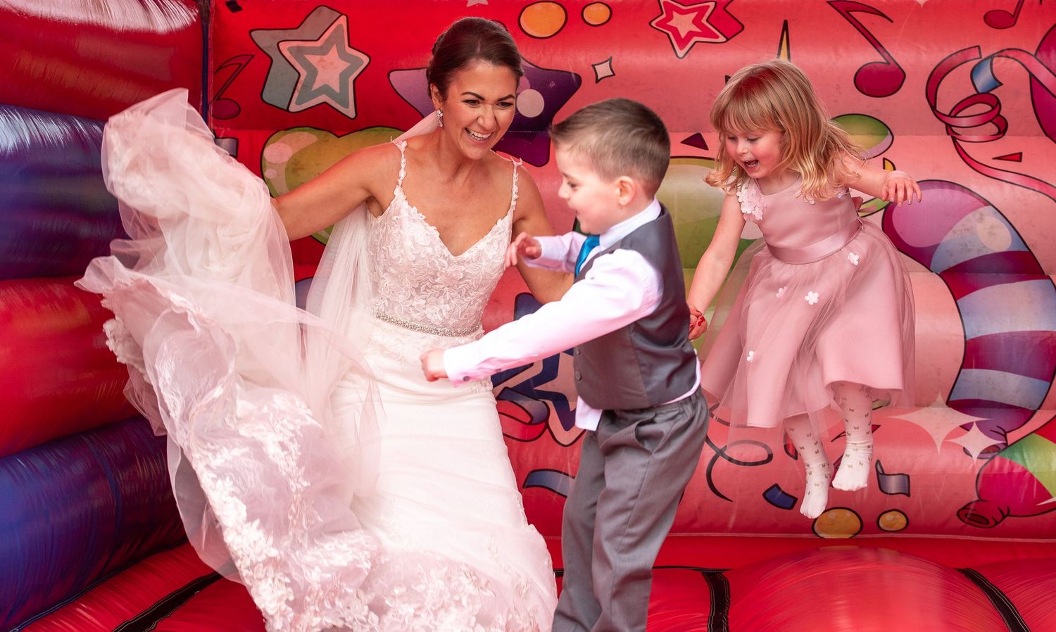 People celebrate on a colorful bouncy castle as formal wear swirls during a festive wedding moment.