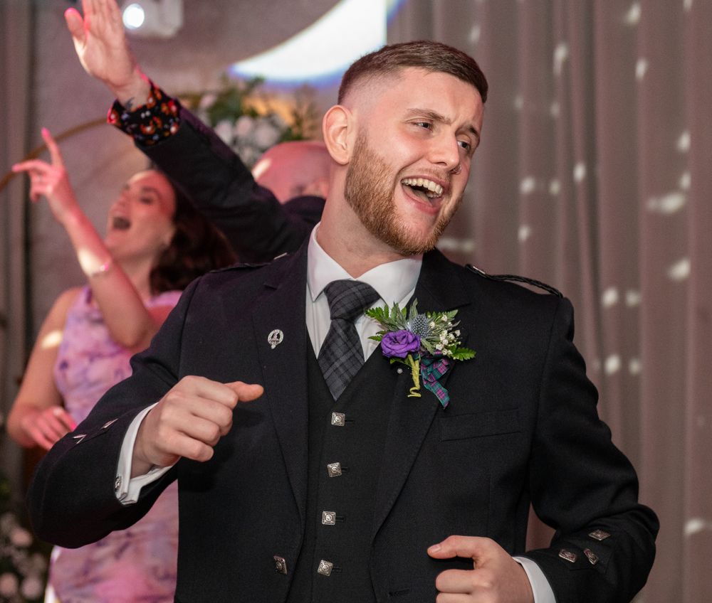A groom in traditional Scottish formal wear dancing and celebrating at a wedding reception.