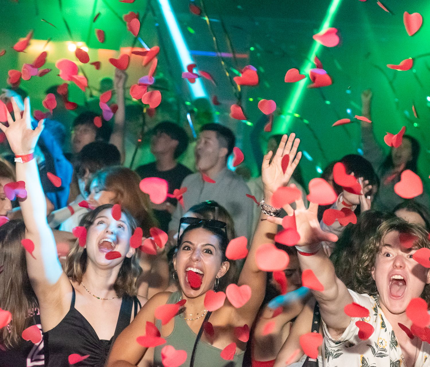Crowd of concert attendees cheering with raised hands as red rose petals fall under green and blue stage lighting.