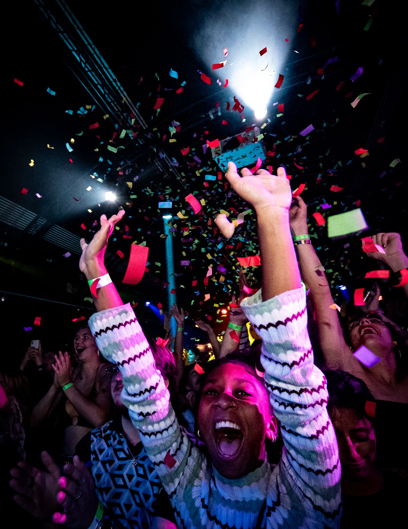Excited crowd celebrating at a nightclub with colorful confetti falling and bright lights illuminating the dark space.