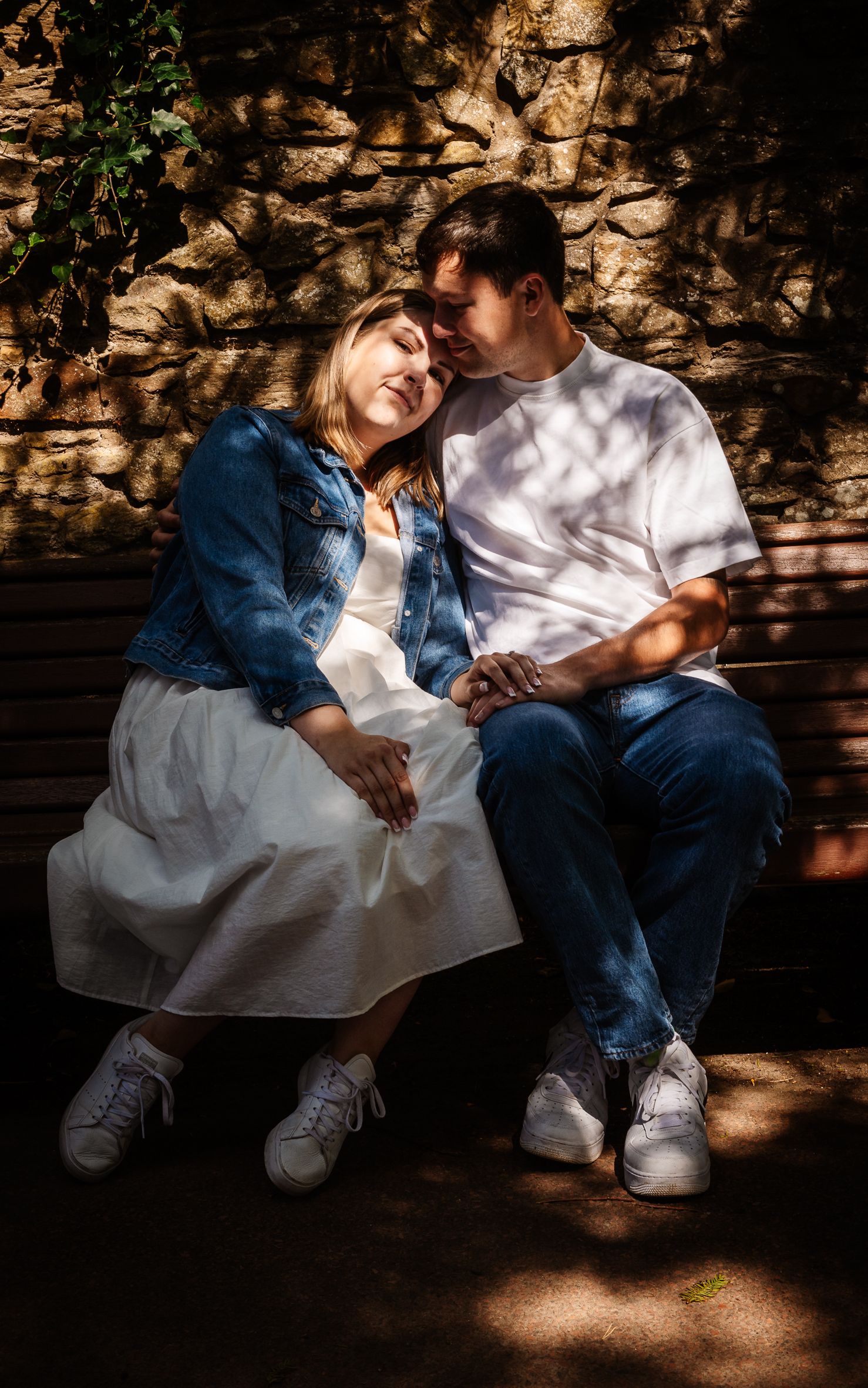 A couple in denim and white sits on a bench in dappled sunlight, sharing a moment together at the royal botanic gardens in Edinburgh