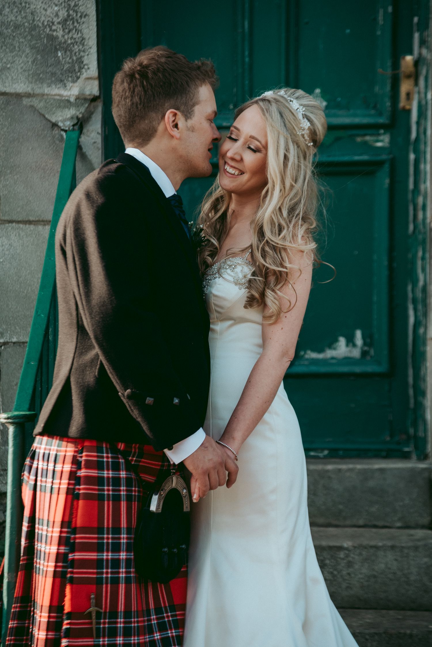 A joyful wedding couple embraces by a green door, with the groom wearing a traditional Scottish kilt.