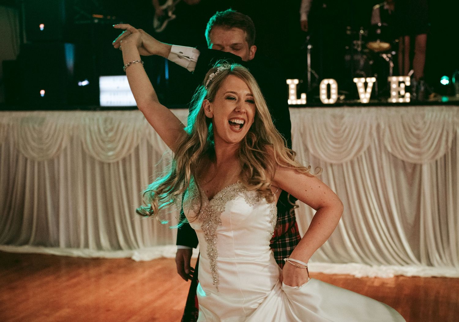 Wedding guests dance and celebrate on a wooden dance floor at an indoor reception venue with white decor and lighting.