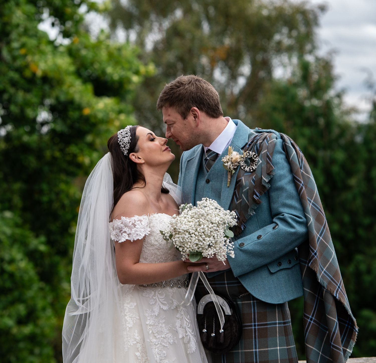 A romantic wedding moment captured outdoors with the groom wearing traditional Scottish kilt and bride holding white bouquet.