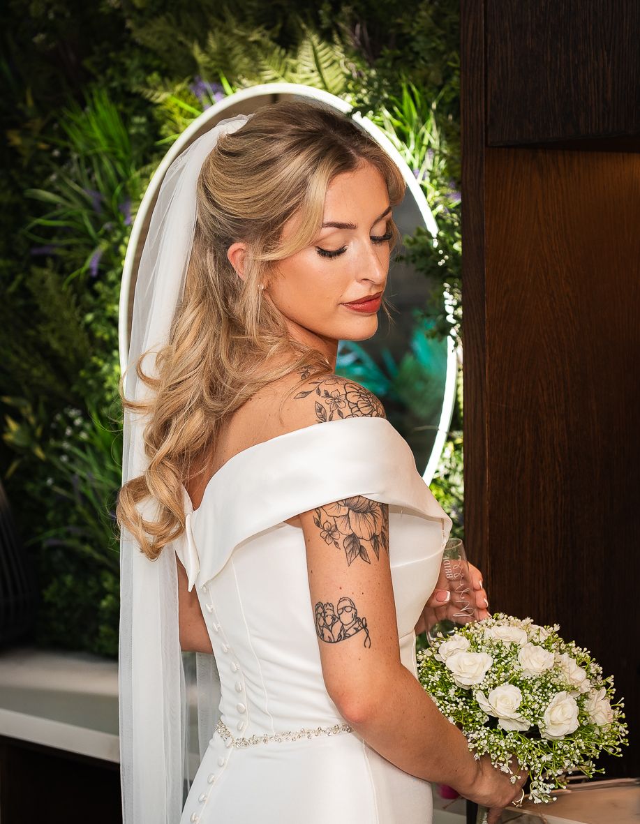 Elegant bride in off-shoulder wedding dress holding white bouquet in garden setting.