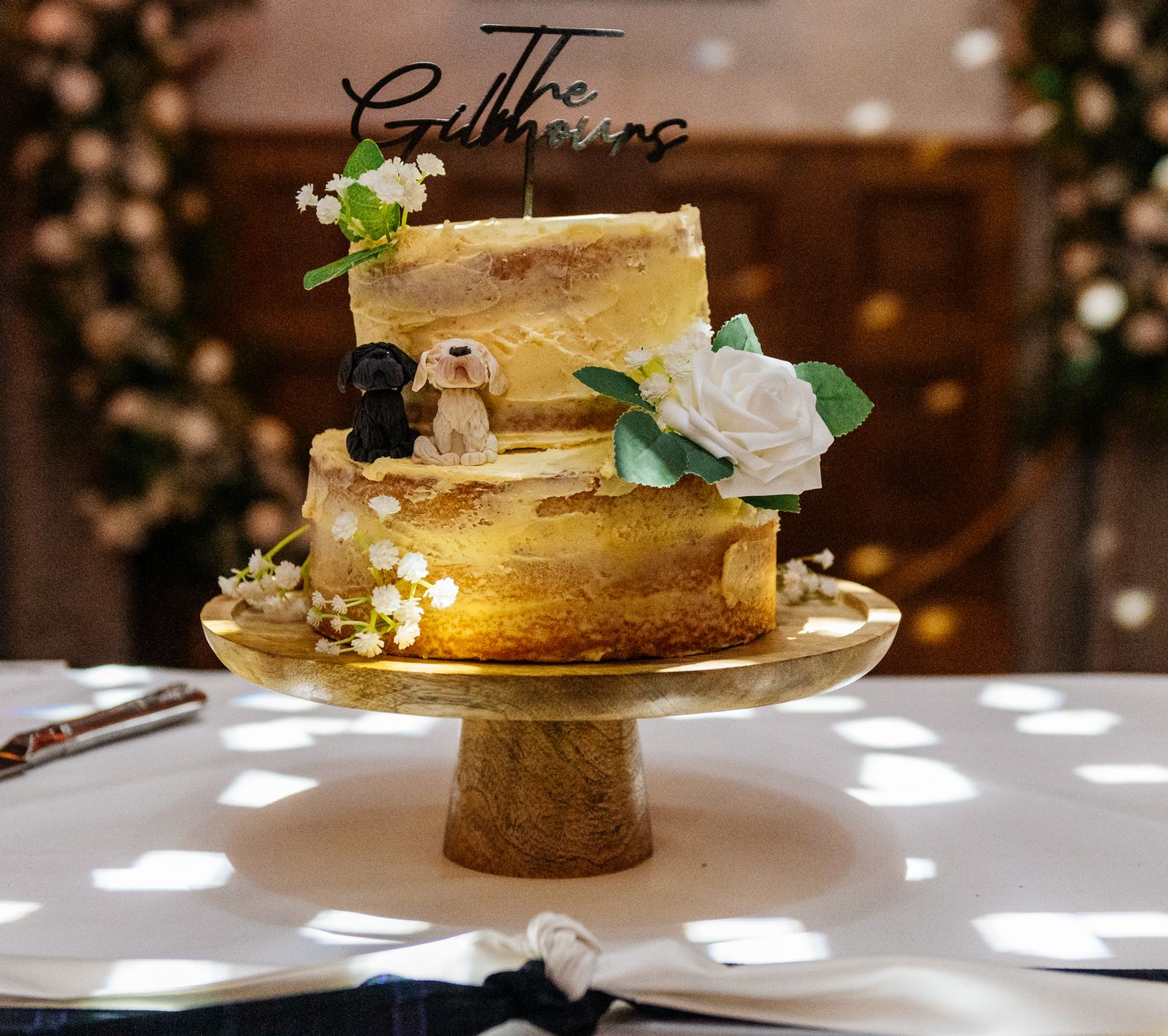 Elegant two-tier gold wedding cake decorated with white roses on wooden stand.