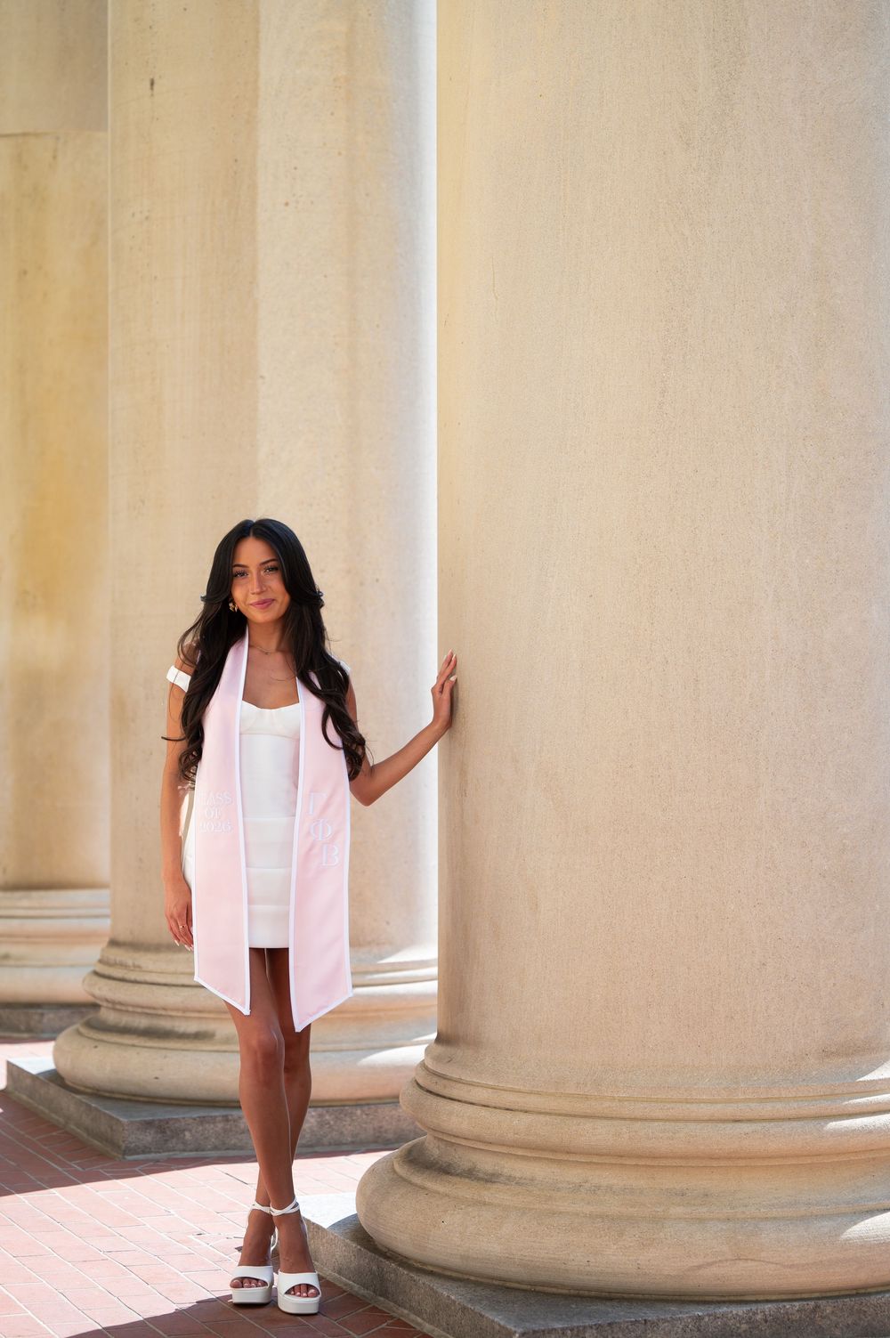 Young woman in white dress and pink vest poses gracefully between large classical columns outdoors.
