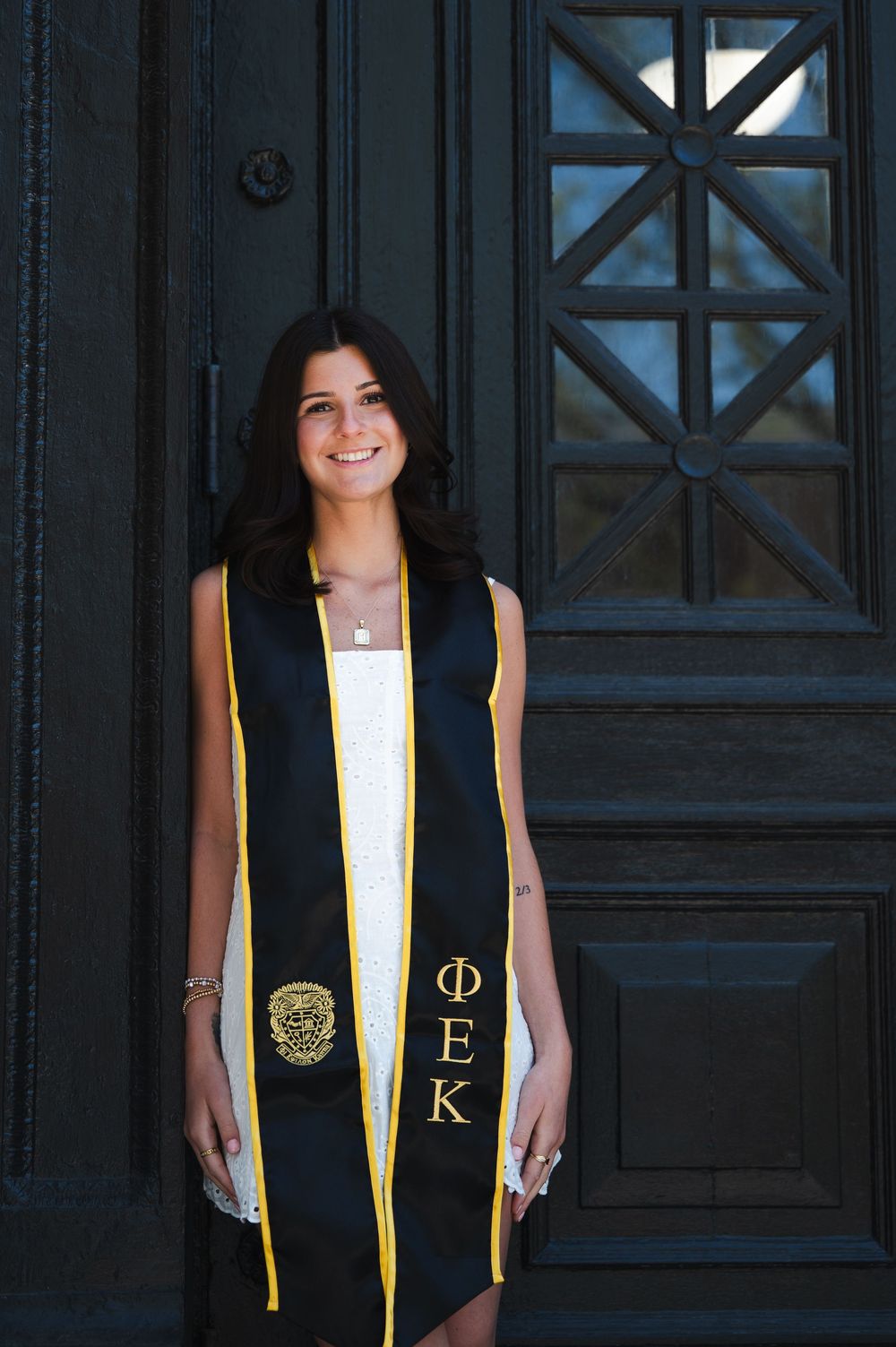 Graduate in black graduation stole with gold trim and Greek letters stands smiling in front of dark wooden door.