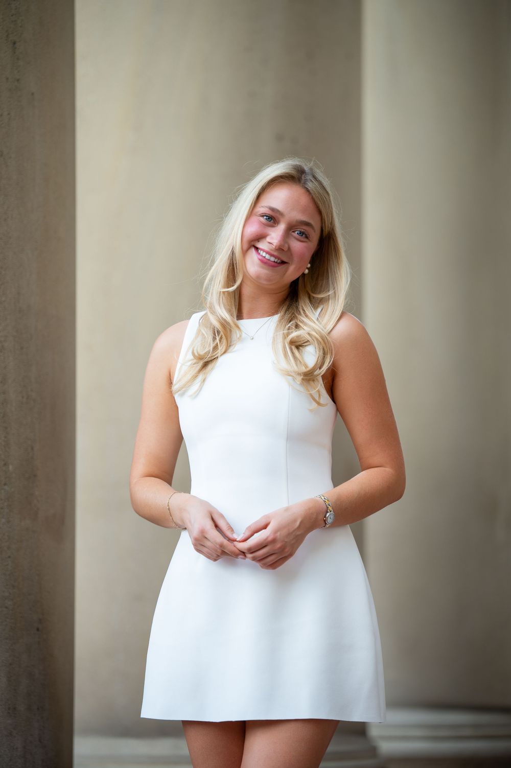 Smiling blonde woman in a white sleeveless mini dress standing between columns outdoors.