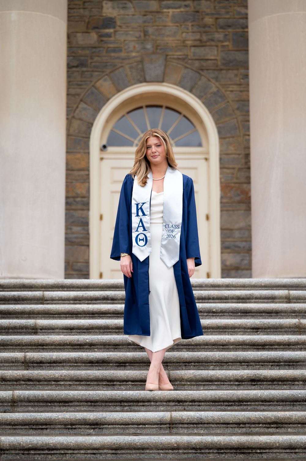 Woman in navy graduation gown with KAΘ sorority stole standing on stone steps before arched doorway.