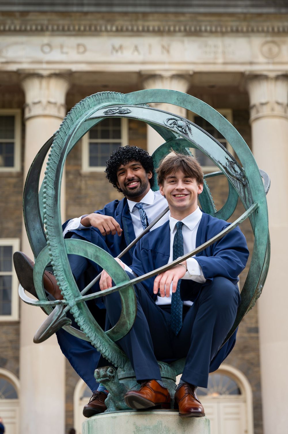 Two graduates in blue robes sit on a bronze armillary sphere sculpture outside Old Main building.