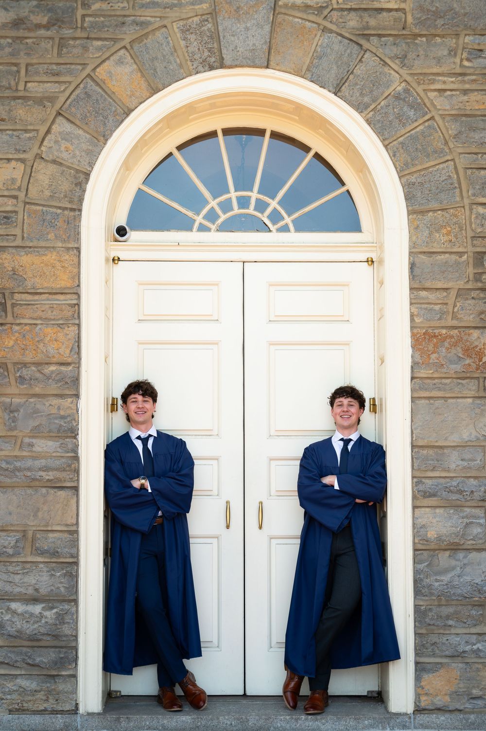 Two young men in blue graduation gowns and ties pose with crossed arms before elegant white arched doors.