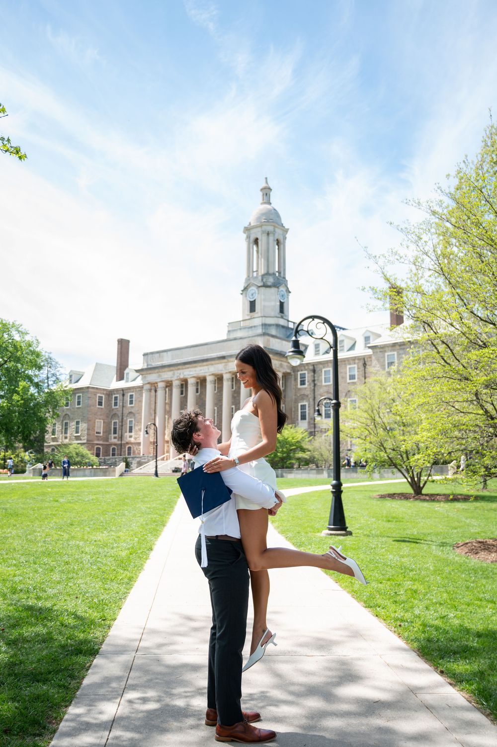 Couple shares romantic lift pose on Penn State campus walkway with Old Main building backdrop on sunny spring day.