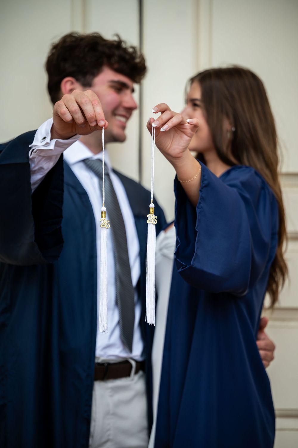 Two graduates in navy blue gowns smiling and holding up their white graduation tassels together.